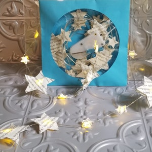 May include: A string of star-shaped fairy lights, crafted from book pages, illuminates a light blue square with a circular cutout. The lights are arranged around the cutout and extend outward, creating a whimsical, decorative effect.