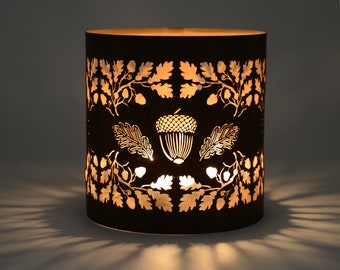 May include: Black metal candle holder with a cut-out design of acorns and oak leaves. The candle holder is lit and casting a warm glow.
