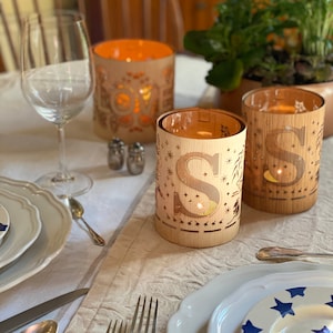 May include: Three wooden candle holders with cut-out designs. The candle holders are lit and have a warm glow. The candle holders are on a white tablecloth with silverware and plates.