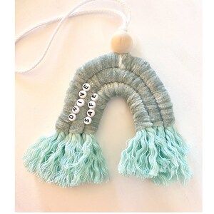 Puede incluir: Un arcoíris de crochet azul claro y blanco con las palabras "DRIVE SAFE" escritas en cuentas blancas. El arcoíris tiene una cuerda blanca unida a una cuenta de madera en la parte superior.