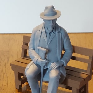 以下が含まれることがあります： 帽子とスーツを着た人物のグレーのフィギュアが、杖を持ったまま木のベンチに座っています。