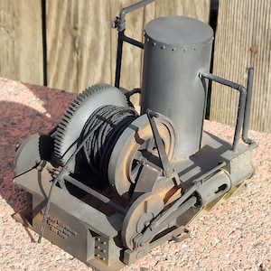 May include: A vintage-style, gray metal winch with a black rope wrapped around the spool. The winch has a metal lever and a small, gray metal tank on top. The winch is labeled "Lady Bess Works, Spokane, WA, Est. 1888".