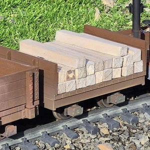 May include: A model train car loaded with wooden beams. The car is brown with metal wheels and sits on a track. The beams are light-colored and stacked neatly. A red car filled with coal is also visible.