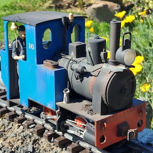 May include: A blue and red toy train with a black boiler and a bell on the roof. The train has a number "16" on the side. The train is on a track and there is a person in the cab.