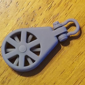 Könnte beinhalten: Eine graue Kunststoffrolle mit einer Metallschlaufe. Die Rolle hat ein Rad mit acht Speichen und ein kleines Loch in der Mitte.