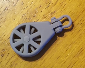 May include: A gray plastic pulley with a metal loop. The pulley has a wheel with eight spokes and a small hole in the center.
