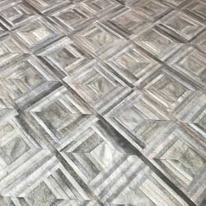 May include: A gray and white geometric patterned rug made of leather. The rug is made up of squares and rectangles of different sizes and colors.