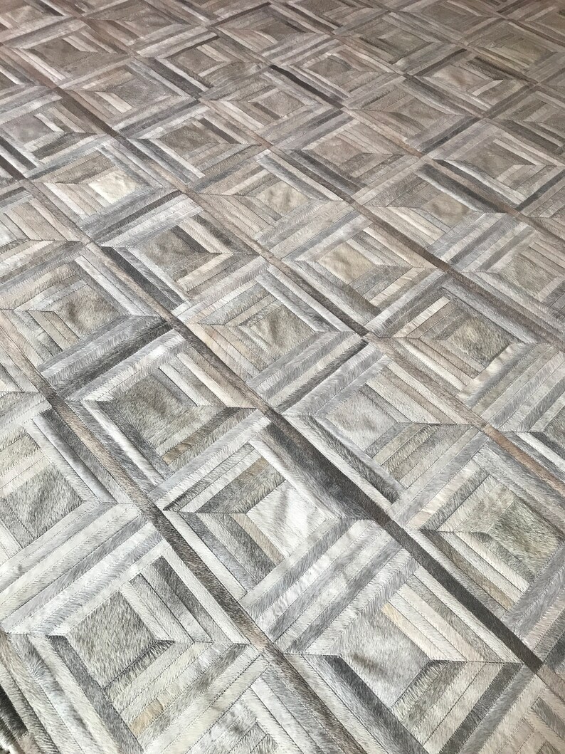 May include: A gray and white geometric patterned rug made of leather. The rug is made up of squares and rectangles of different sizes and colors.