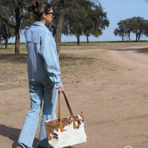 May include: A woman wearing a blue denim jacket and jeans walks along a dirt path, carrying a white and brown cowhide tote bag with brown leather handles.