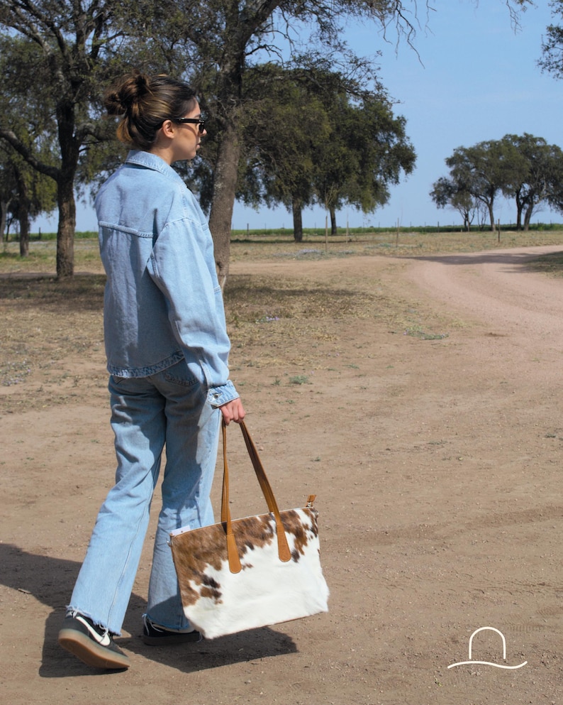 May include: A woman wearing a blue denim jacket and jeans walks along a dirt path, carrying a white and brown cowhide tote bag with brown leather handles.