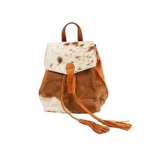May include: A brown and white cowhide leather backpack with a flap closure and tassel accents.