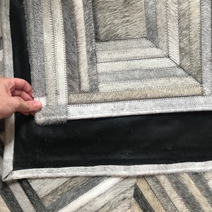 May include: A gray and white cowhide rug with a geometric pattern. The rug is made of different pieces of cowhide that have been sewn together. The rug has a black backing.