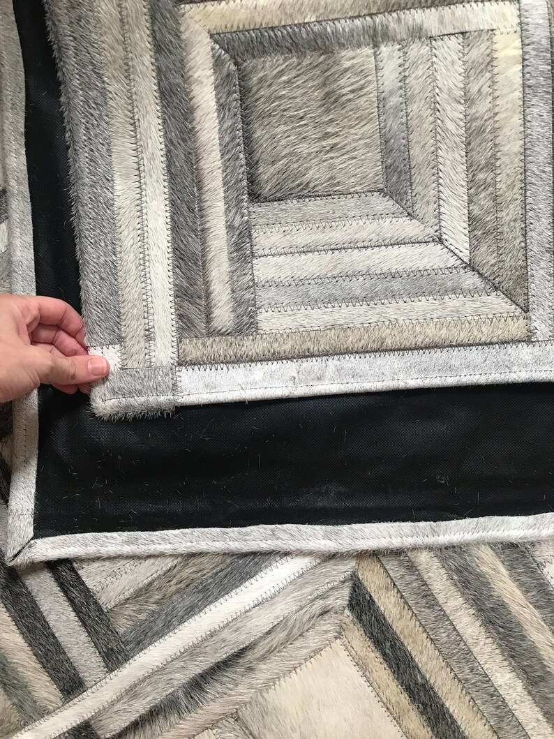 May include: A gray and white cowhide rug with a geometric pattern. The rug is made of different pieces of cowhide that have been sewn together. The rug has a black backing.