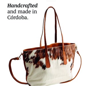 May include: A brown leather tote bag with a cowhide pattern. The bag has two top handles and a long shoulder strap. The bag is handcrafted and made in Cordoba.
