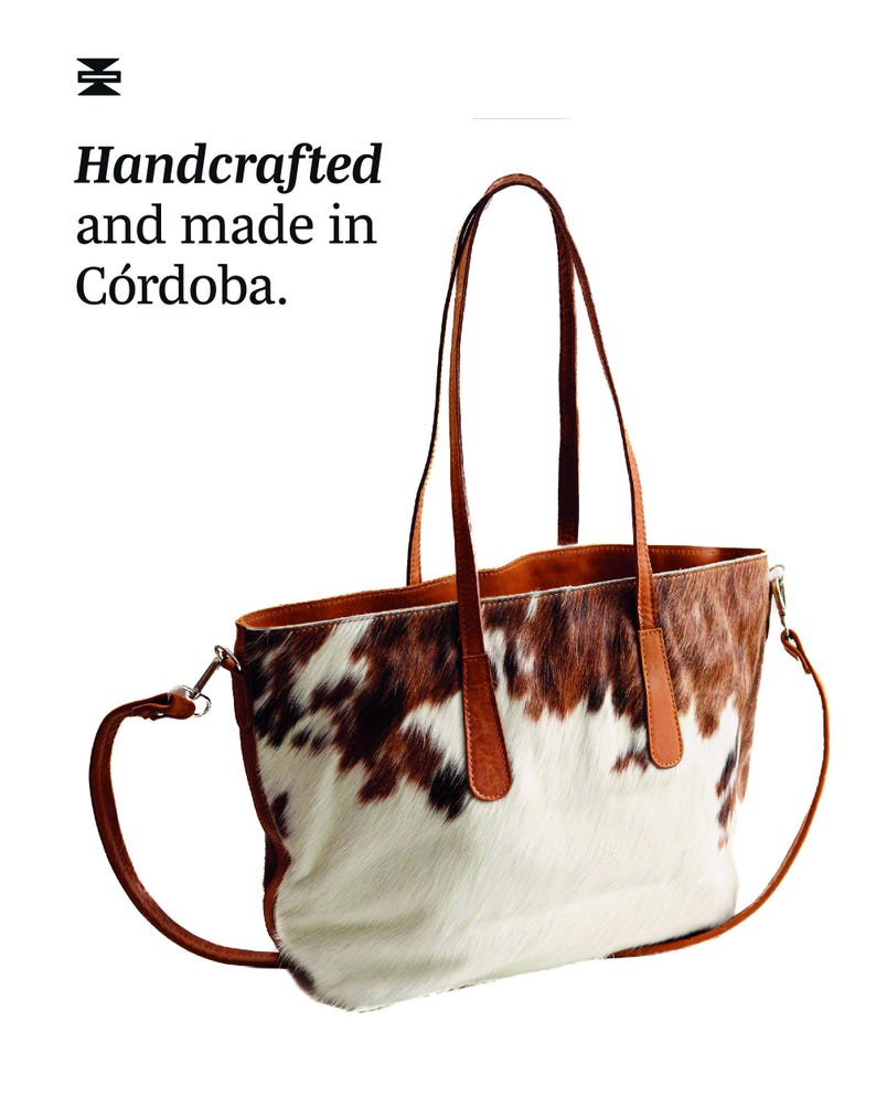 May include: A brown leather tote bag with a cowhide pattern. The bag has two top handles and a long shoulder strap. The bag is handcrafted and made in Cordoba.