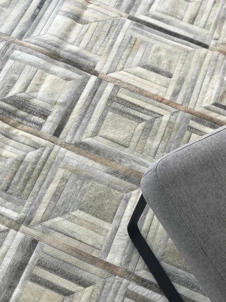 May include: A gray and white patchwork rug with a geometric pattern. The rug is made of different pieces of leather sewn together. A gray chair with black legs is partially visible in the bottom right corner of the image.