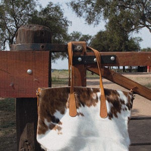 May include: A brown and white cowhide tote bag with brown leather handles hanging on a wooden fence.