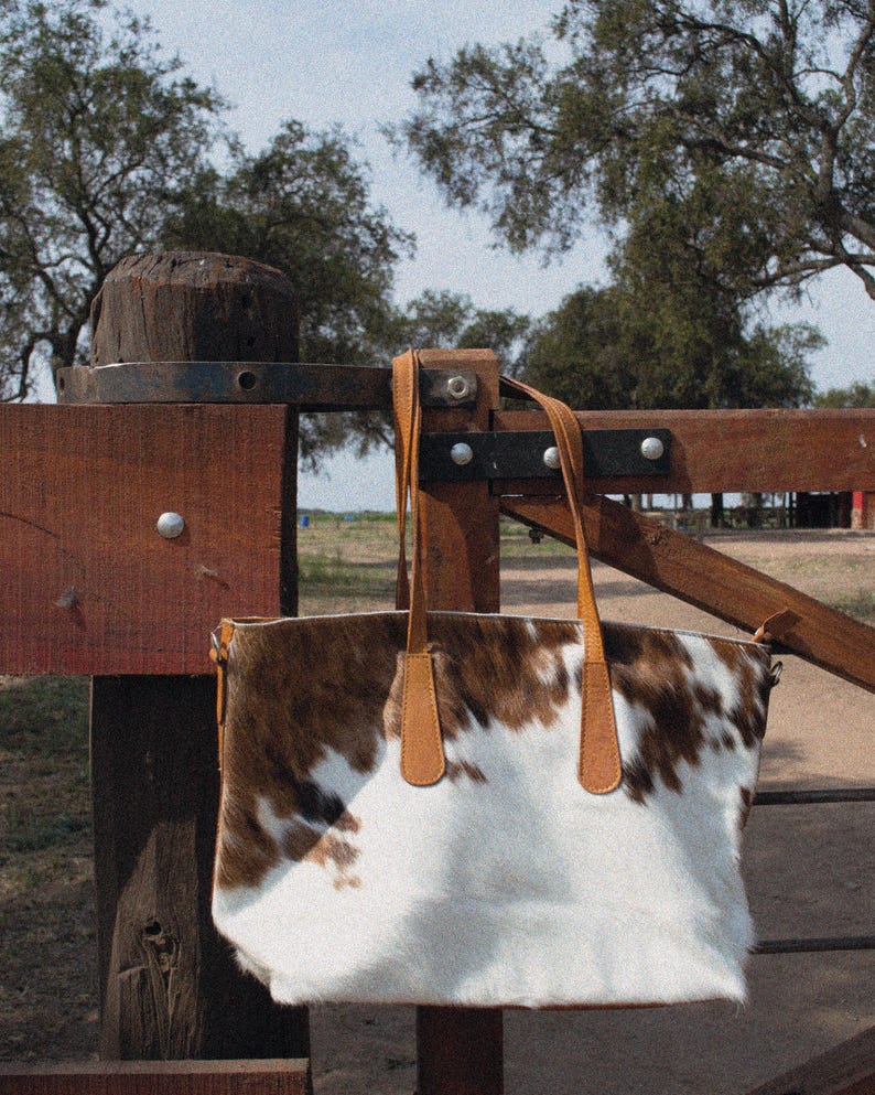 May include: A brown and white cowhide tote bag with brown leather handles hanging on a wooden fence.