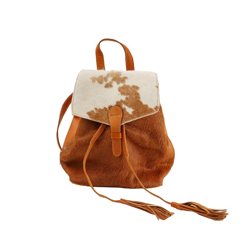 May include: A brown leather backpack with a white and brown cowhide flap. The backpack has a drawstring closure and two long tassels.