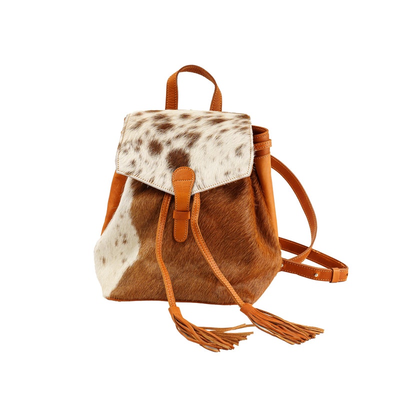 May include: A brown leather backpack with a white and brown cowhide pattern. The backpack has a flap closure with a leather strap and tassel accents.
