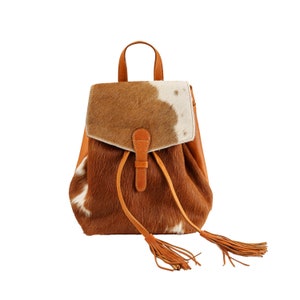 May include: A brown leather backpack with a cowhide pattern and fringe tassels. The backpack has a flap closure with a buckle and a drawstring closure.