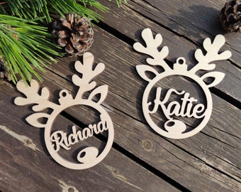 May include: Two wooden reindeer ornaments with the names "Richard" and "Katie" cut out in a cursive font. The ornaments have antlers and a circular frame. The background includes pine needles and pine cones.