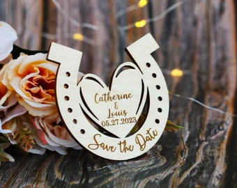 May include: Wooden horseshoe save the date magnet with a heart cutout and the text "Catherine & Louis 05.27.2023 Save the Date".