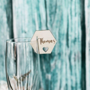 Può includere: Un segnaposto esagonale in legno con il nome "Thomas" scritto in lettere dorate. Un piccolo cuore è tagliato dalla carta.