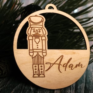 Può includere: Ornamento natalizio in legno con un design schiaccianoci tagliato al laser all'interno di una cornice circolare. Il nome "Adam" è scritto sotto. L'ornamento è in legno chiaro e pronto per essere appeso.