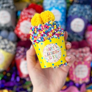 May include: A cupcake-shaped sock with a yellow paper cup, blue polka dots, and a "Happy Birthday To You" sticker. The sock has colorful sprinkles and two yellow pom-poms on top. Other cupcake socks are in the background.