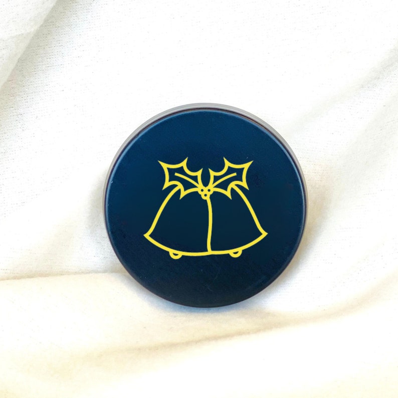 May include: A dark blue round container with a yellow design of two bells with holly leaves and berries.