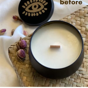 May include: A black candle with a white wax top and a wooden wick. The candle is in a black tin container with a lid that has a gold eye design. The word "before" is written on a white surface behind the candle.