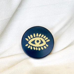 May include: A black round coaster with a gold eye design. The eye is stylized with a yellow iris and pupil and a black outline.