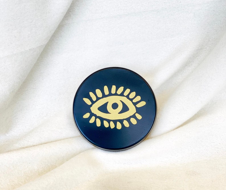 May include: A black round coaster with a gold eye design. The eye is stylized with a yellow iris and pupil and a black outline.