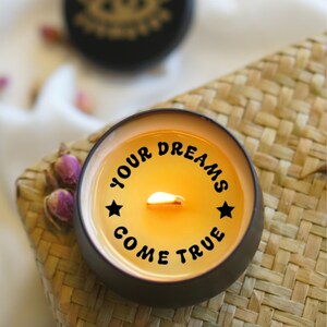 May include: A black candle with a yellow wax interior and a lit wick. The candle has the text "YOUR DREAMS COME TRUE" printed around the side in black.