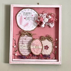 Op de afbeelding: Een roze omlijste Paas-schaduwdoos. De doos heeft een rond bord met "Happy Easter" en konijnenoren, drie decoratieve eieren en een stoffen bloem. De eieren zijn versierd met kant, lint en touw. Paarse bloemen en groen bevinden zich aan de basis.