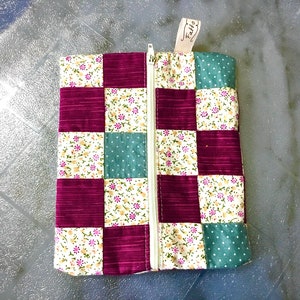 Può includere: Una pochette con cerniera con un design a patchwork che presenta quadrati di colore bordeaux, bianco con piccola stampa floreale e verde acqua con pois bianchi. La pochette ha una cerniera bianca e un'etichetta bianca che dice "Fatto".