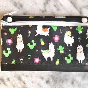 May include: Black zippered pouch with a white zipper and white zipper pulls. The pouch has a repeating pattern of llamas, cacti, pineapples, and flowers on a black background.
