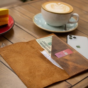 May include: A brown leather wallet with a credit card and cash inside. The wallet is open and lying on a wooden table with a cup of coffee and a piece of cake in the background.