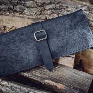 May include: A black leather case with a silver buckle and a strap. The case is lying on a pile of wooden logs.