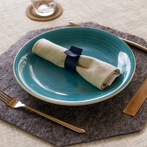 May include: A blue ceramic dinner plate with a white rim sits on a gray felt placemat. A white linen napkin is rolled and tied with a blue ribbon. A silver fork and a silver knife are on the placemat.