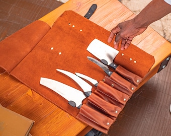 May include: A brown leather knife roll with six slots for knives. The roll is open and the knives are visible. The knives are all different sizes and shapes. The roll is on a wooden table.