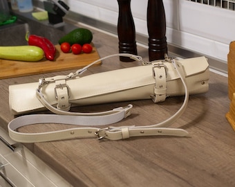 May include: A white leather knife roll with a strap and buckle closure. The roll is lying on a wooden surface.