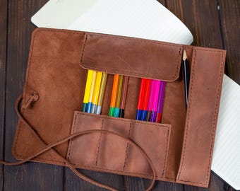 May include: A brown leather pencil roll with a flap closure. The roll is open and filled with colored pencils. There is a black pencil in a separate pocket.
