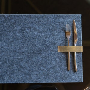 May include: A gray felt placemat with a gold metal utensil holder and a fork and knife placed inside.