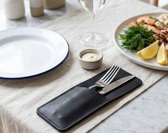 May include: A black leather cutlery holder containing a stainless steel fork and knife, resting on a beige linen table runner. The holder features visible stitching along the edges and a smooth, matte finish, providing a sleek storage solution for dining utensils.