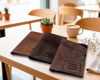 May include: Three rectangular leather menu covers in shades of brown and black, each featuring a square embossed area with the text YOUR LOGO HERE. The covers are arranged in a row on a light wood surface, showcasing a smooth, matte texture.