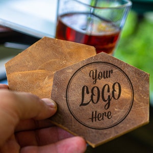 Könnte beinhalten: Zwei braune Lederuntersetzer in sechseckiger Form. Der obere Untersetzer hat den Text "Your Logo Here" in einem Kreis eingraviert.