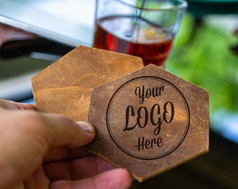 May include: Two brown leather coasters with a hexagonal shape. The top coaster has the text "Your Logo Here" engraved in a circle.