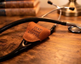 May include: A black medical stethoscope featuring a tan leather cover attached to the tubing. The leather cover is triangular in shape and has the text "STAT! I need COFFEE." embossed in black lettering on its front surface.